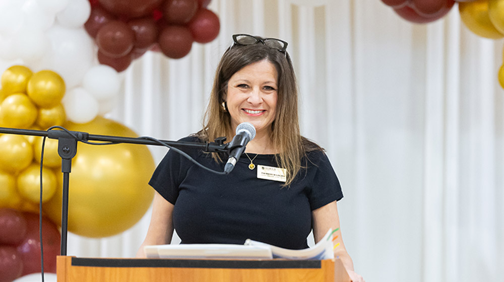Tina Marino-Misura standing in behind a microphone at a podium and smiling